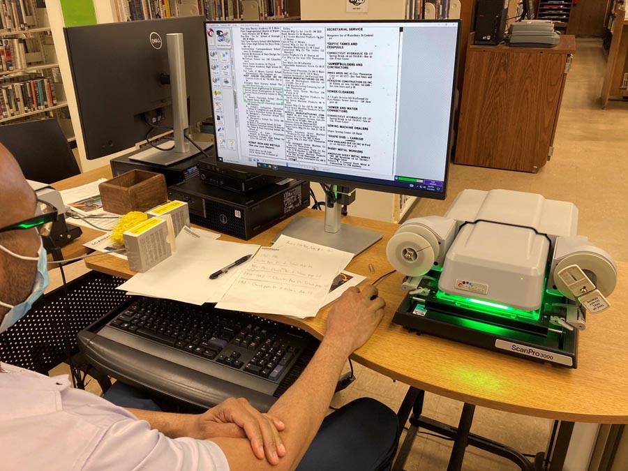 Librarian sitting at the microfilm computer station reading through a scan of a historical newspaper.